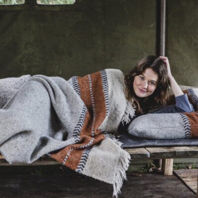 Eine Frau ruht sich auf einem Holzbett in einem grünen Zelt aus und lächelt in die Kamera, während sie ihren Kopf auf ein Kissen stützt und sich mit der gestreiften Montana Grey Leinen-Wolle-Mix von Libeco-Decke zudeckt.