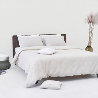 A minimalist bedroom with a neatly made bed in Natural Classic Linen Bedding by Geniksa, two pillows, a white basket with cushions, books on the floor, tall pampas grass in a vase and calming neutral tones.