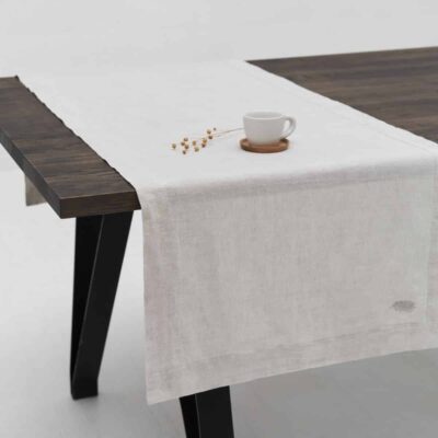 A dark wooden table with the Natural linen tablecloth by Geniksa as a light beige runner, paired with a white cup and saucer and a small dried plant. The minimalist white background underscores this elegant ambience.