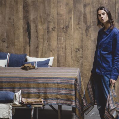 Eine Person in einer blauen Jacke steht an einem Tisch, der mit Olympia Stripe - reines Leinen für den Tisch von Libeco gedeckt ist, und hält ein passendes Tuch. Holzwände und Kissen auf einer nahen Bank vervollständigen die Szene.