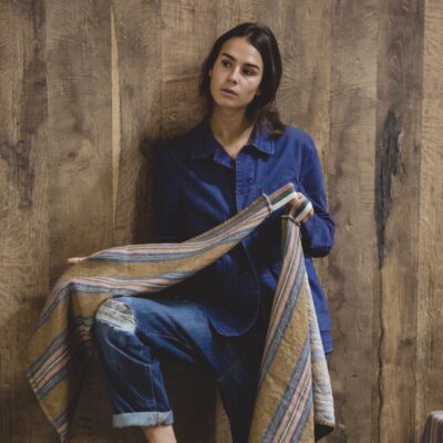 Eine Frau in blauem Jeanshemd und Jeans steht an einer Holzwand, hält das Olympia - reines Leinenhandtuch von Libeco in der Hand und blickt nachdenklich zur Seite.