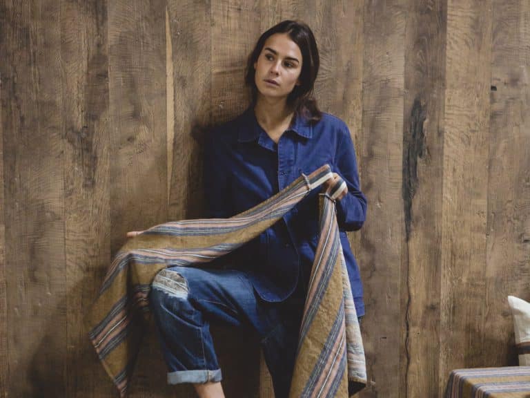 Eine Frau in blauem Jeanshemd und Jeans steht an einer Holzwand, hält das Olympia - reines Leinenhandtuch von Libeco in der Hand und blickt nachdenklich zur Seite.