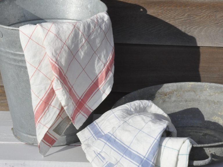 Zwei Metallwaschwannen stehen im Freien; in der einen ist ein weißes Tuch mit roten Streifen, in der anderen ein weißes Tuch mit blauen Streifen drapiert.