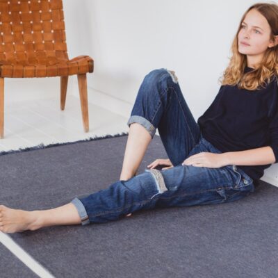 Eine Frau mit langen Haaren, die einen dunklen Pullover und zerrissene Jeans trägt, sitzt barfuß auf einem dunklen Teppich in einem hellen, minimalistischen Raum mit einem geflochtenen Ledersessel im Hintergrund.
