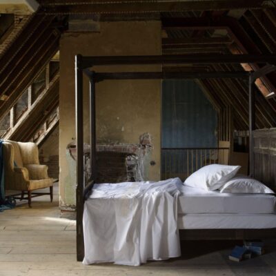 Das rustikale Dachgeschoss-Schlafzimmer verfügt über ein hölzernes Himmelbett, das mit Prager Percal 400TC Bettwäsche von Peter Reed bezogen ist. Ein blauer Sessel neben freiliegenden Balken und einem abblätternden Kamin vervollständigen den Vintage-Charme des Zimmers.