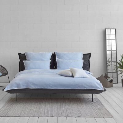 Ein modernes Schlafzimmer mit einem hellblauen Bett, einem schwarzen Kopfteil, zwei Kissen, einem schwarzen Drahtstuhl, einem hohen Spiegel, einer Topfpflanze und einem kleinen grauen Teppich auf weißem Holzboden vor einer weißen Backsteinwand.