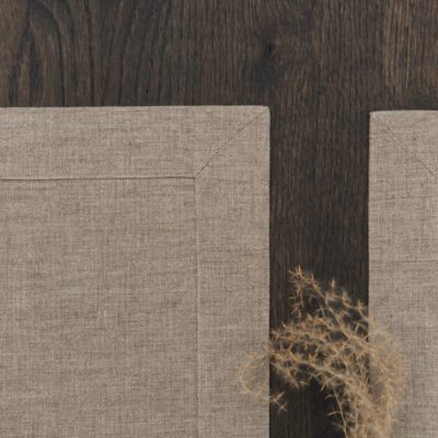 Zwei beigefarbene Leinentischsets auf einem dunklen Holztisch, mit getrocknetem Gras oder Laub, das in der rechten unteren Ecke teilweise sichtbar ist. Die Tischsets sind sauber platziert und haben sichtbare Ecknähte.