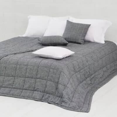 A neatly made bed with Scotia Classic Linen Bedding by Geniksa – featuring a grey quilted bedspread, grey and white pillows – stands against white walls and floor. A black side table and lamp add a modern touch.