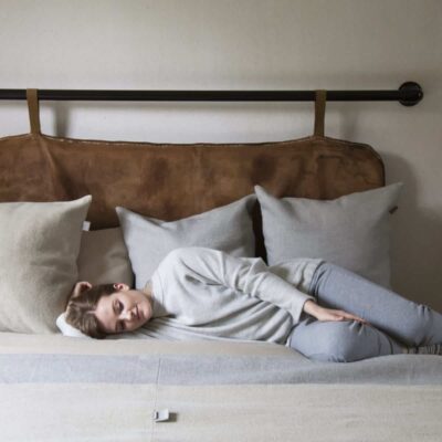 Eine Person mit kurzen Haaren in einem grauen Outfit liegt auf der Seite auf einem Bett mit beigen und grauen Kissen und stützt ihren Kopf auf ein Kissen in einem gemütlichen, minimalistischen Schlafzimmer.