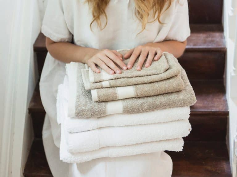 Eine Frau in einem weißen Kleid sitzt auf einer Holztreppe und hält einen Stapel Simi Leinenfrottier fürs Bad Flax von Libeco in der Hand. Ihre Hände ruhen sanft auf der sauber gefalteten weißen und beigen Wäsche.