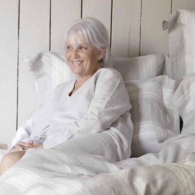 Eine ältere Frau mit grauem Haar und einem hellen Oberteil sitzt auf einem Bett mit gestreifter Bettwäsche, lächelt und entspannt sich vor dem Hintergrund einer Holzwand.