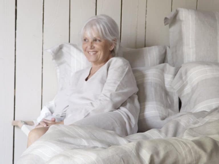 An older woman with gray hair and a light-colored top sits on a bed with striped bed linen, smiling and relaxing against the backdrop of a wooden wall.