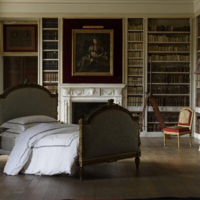 Ein großes Schlafzimmer mit zwei antiken Betten, bezogen mit Stave Percal 400TC Bettwäsche von Peter Reed, weißem Bettzeug, Bücherregalen an den Wänden, einem Kamin mit einem Porträt, einem kleinen roten Stuhl und einer Stehlampe mit einem bunten Schirm.