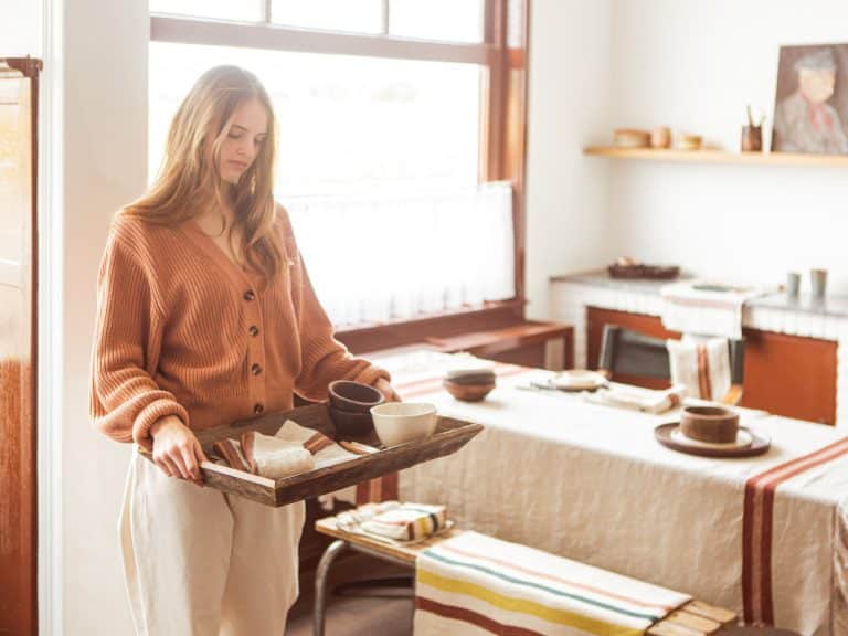A woman in a cozy, sunlit dining room carries a wooden tray with cups and bowls. The table is set with a striped tablecloth and simple, rustic crockery, creating a warm, inviting atmosphere.