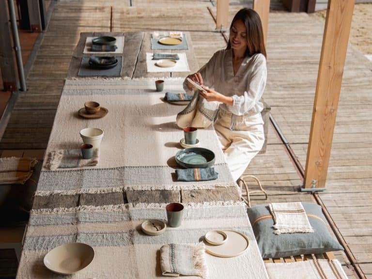 Eine Frau faltet eine Stoffserviette an einem langen, mit Tellern, Tassen und Schüsseln gedeckten Tisch auf einer Holzterrasse. Der Tisch ist mit der belgischen Tischdecke Ioulida reines Leinen von Libeco drapiert, während natürliches Licht einfällt.