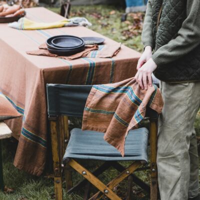 Eine Person steht im Freien an einem mit Tellern und Servietten gedeckten Tisch inmitten von Herbstblättern und hält The Ontario Stripe - reines Leinenhandtuch von Libeco über einen Stuhl neben dem mit einem braun gestreiften Tuch bedeckten Tisch.
