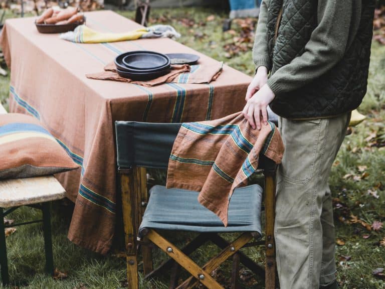 Eine Person steht im Freien an einem mit Tellern und Servietten gedeckten Tisch inmitten von Herbstblättern und hält The Ontario Stripe - reines Leinenhandtuch von Libeco über einen Stuhl neben dem mit einem braun gestreiften Tuch bedeckten Tisch.