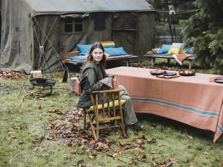 A woman sits at an outdoor table covered with a striped cloth, surrounded by autumn leaves. Behind her are benches with colorful cushions and a large green tent, suggesting a cozy outdoor gathering or campsite.