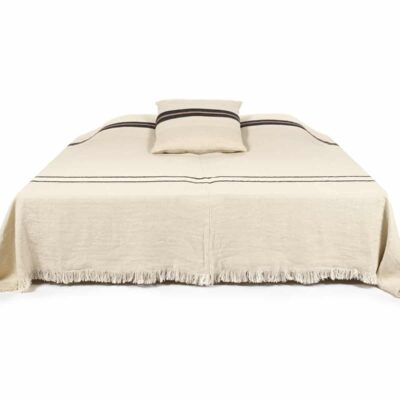 Ein beigefarbenes Bett mit fransenbesetzter Tagesdecke und passendem Kissen, beide mit schwarzen Querstreifen, steht vor einem weißen Hintergrund.