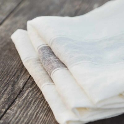 Drei fein säuberlich gefaltete Ajaccio - reines Leinen für die Küche von Libeco Servietten in Weiß und Off-White werden auf einer rustikalen Holzoberfläche präsentiert, die die Texturen von Leinen und Holz hervorhebt.