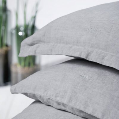 Two neatly stacked light grey linen pillows with subtle embroidery rest in front of tall green plants – the perfect complement to the Caroline Classic Linen Bedding by Geniksa.
