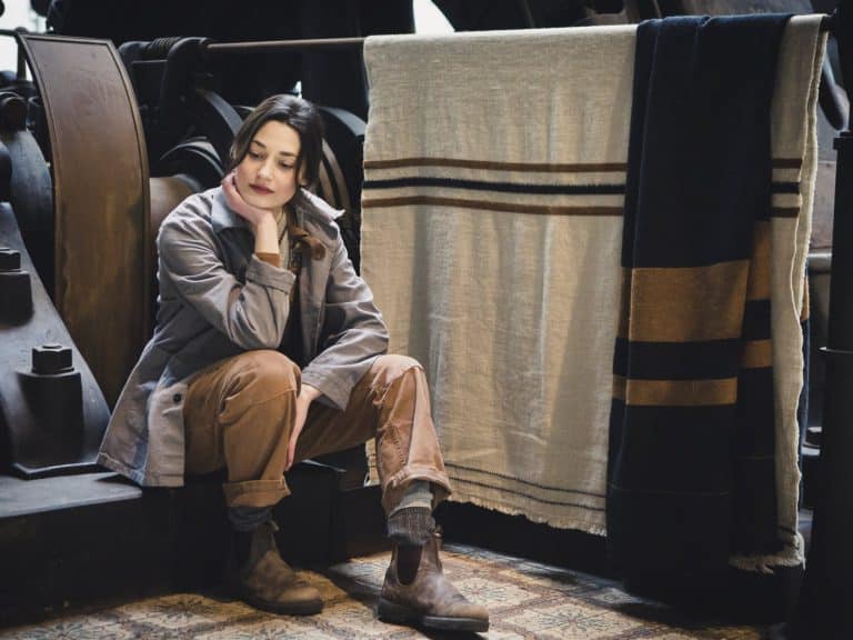 A woman in casual work clothes sits on the floor, her chin resting on her hand, next to large machines and hanging blankets with brown and cream-colored stripes. She looks thoughtful and relaxed.