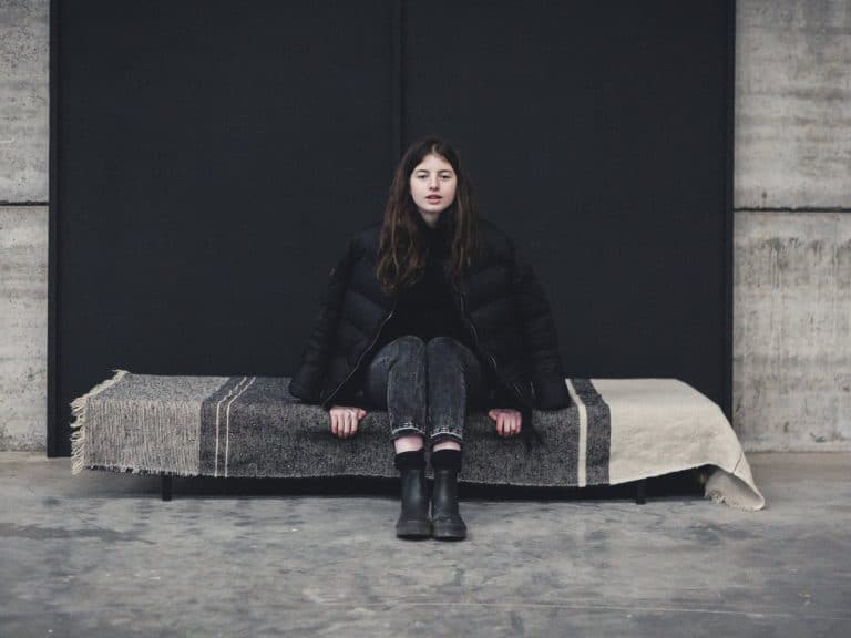 A woman with long brown hair sits on a bench covered with a striped blanket. She is wearing a black puffer jacket, black jeans and black boots against a modern backdrop of concrete and a black wall.