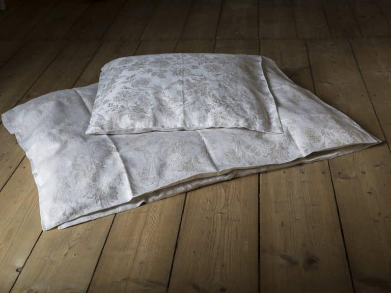 A patterned white cushion and a white blanket lie on a wooden floor, with the soft natural light illuminating the bedding and highlighting the texture of the wooden floorboards.