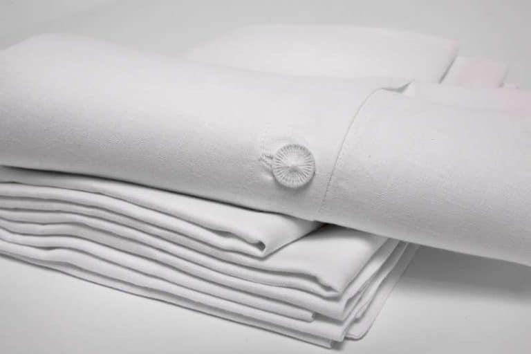 Ein ordentlich gefalteter Stapel Weiße Leinen Bettwäsche mit Hohlsaum von Hoffmann, mit einer dekorativen Knopfverzierung auf einem Stück, ruht auf einer schlichten weißen Oberfläche.