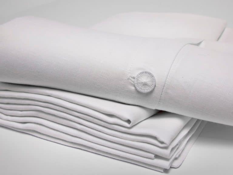 Abgebildet ist ein ordentlich gefalteter Stapel Starkes Leinen - Weiße Bettwäsche der Leinenweberei Hoffmann mit einer Nahaufnahme der geknöpften Manschette auf dem obersten Blatt vor einem schlichten weißen Hintergrund.