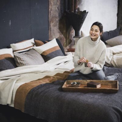 Eine Frau in einem weißen Rollkragenpullover sitzt auf einem Bett mit Nash Leinen-Wolle-Mix von Libeco und hält lächelnd eine Tasse in der Hand. Ein Holztablett mit Snacks und einer weiteren Tasse trägt zur gemütlichen, rustikalen Atmosphäre des Bettes bei.