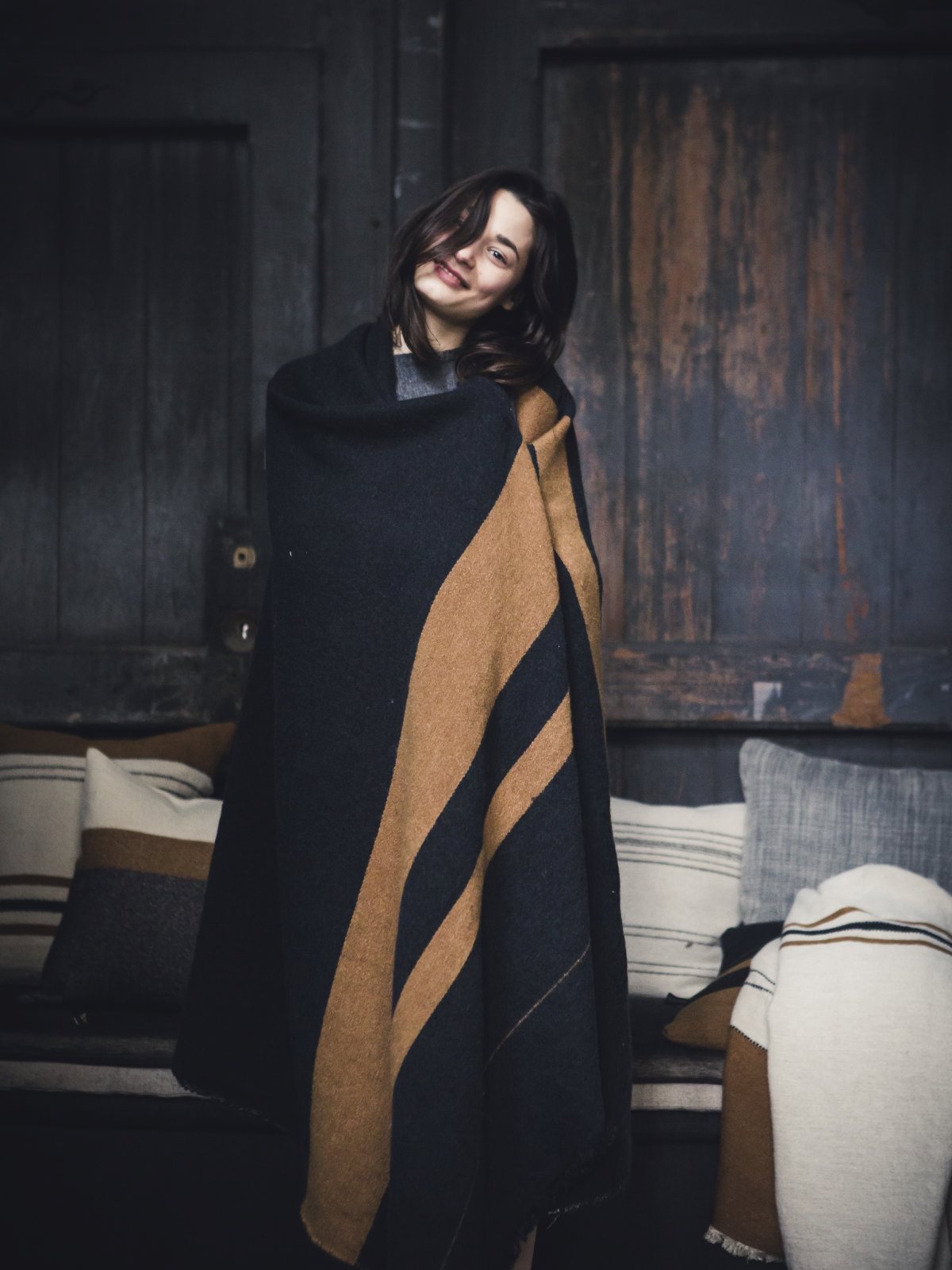 Eine Frau mit langen dunklen Haaren lächelt, während sie in die Decke Oscar Leinen-Wolle-Mix von Libeco gehüllt in einem gemütlichen Raum mit gestreiften Kissen und dunklen Holzwänden steht.