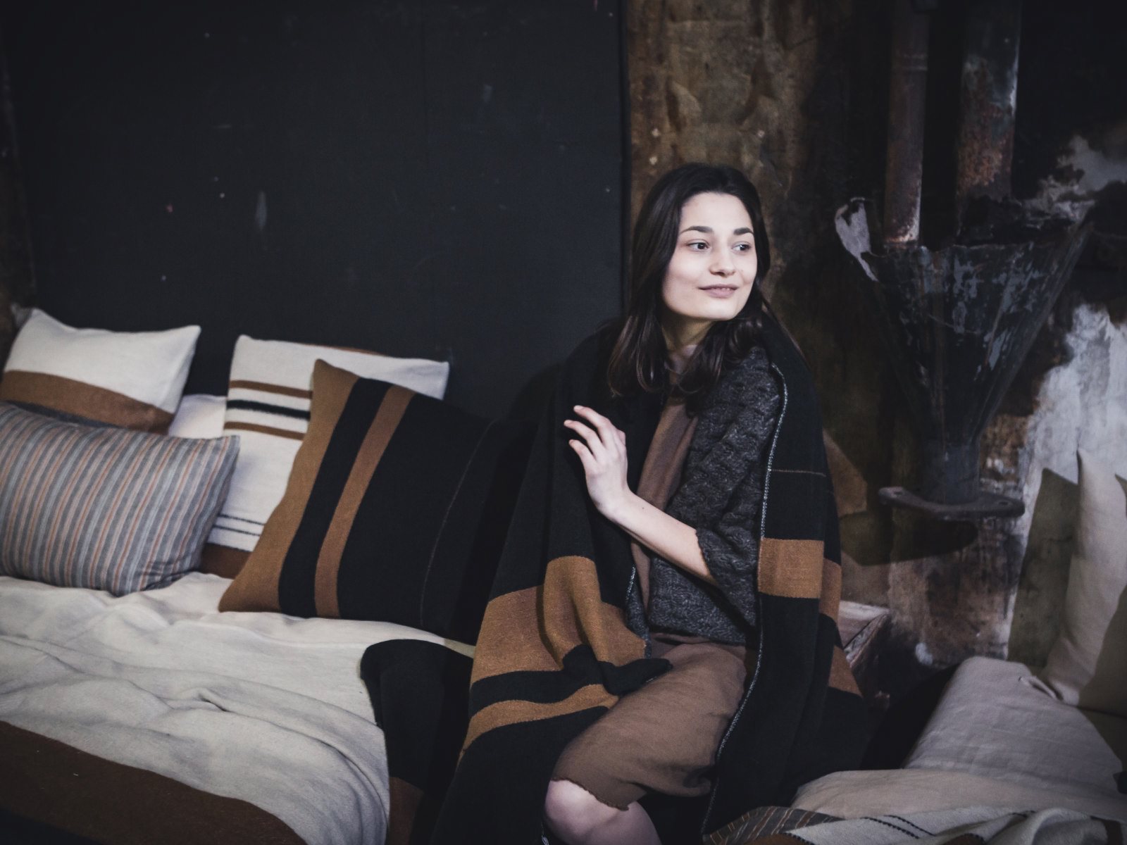 Eine sanft lächelnde Frau sitzt auf einem Bett mit gestreiften Kissen und neutraler Bettwäsche, eingewickelt in die Oscar Leinen-Wolle-Mix von Libeco-Decke mit braunen Streifen, vor einem rustikalen, sanft beleuchteten Hintergrund.