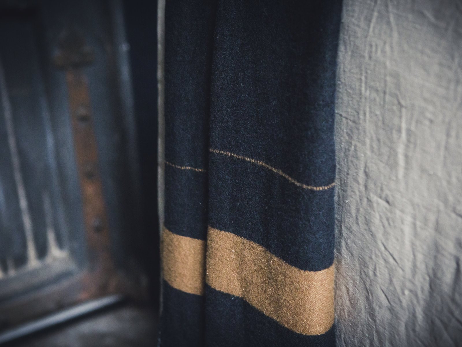 Eine Nahaufnahme des Schals Oscar Leinen-Wolle-Mix von Libeco aus dunkler Wolle mit einem hellbraunen Streifen, der vor einer strukturierten beigen Wand und einem Teil einer rustikalen Tür im Hintergrund hängt.