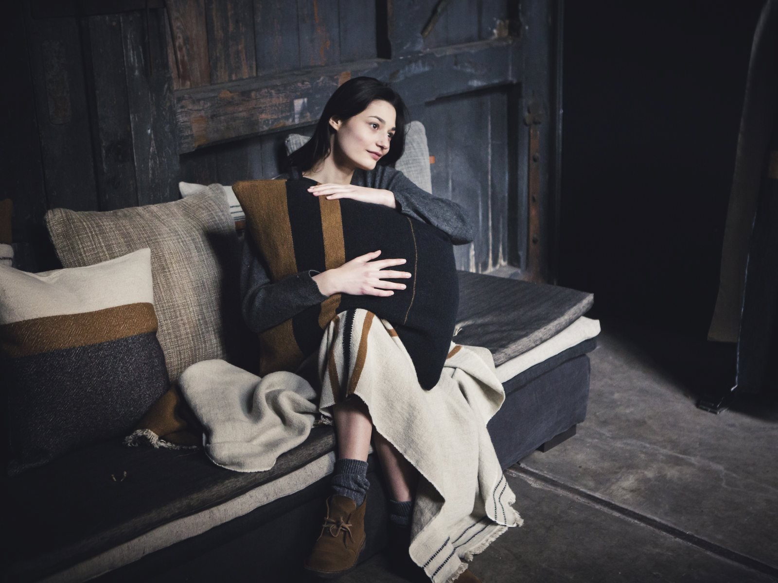 Eine Frau sitzt auf einem Sofa in einem schwach beleuchteten Raum und schmiegt sich an ein Oscar Leinen-Wolle-Mix von Libeco-Kissen. Eingehüllt in eine Decke mit kuscheligen Socken und Stiefeln, blickt sie nachdenklich in die Ferne.