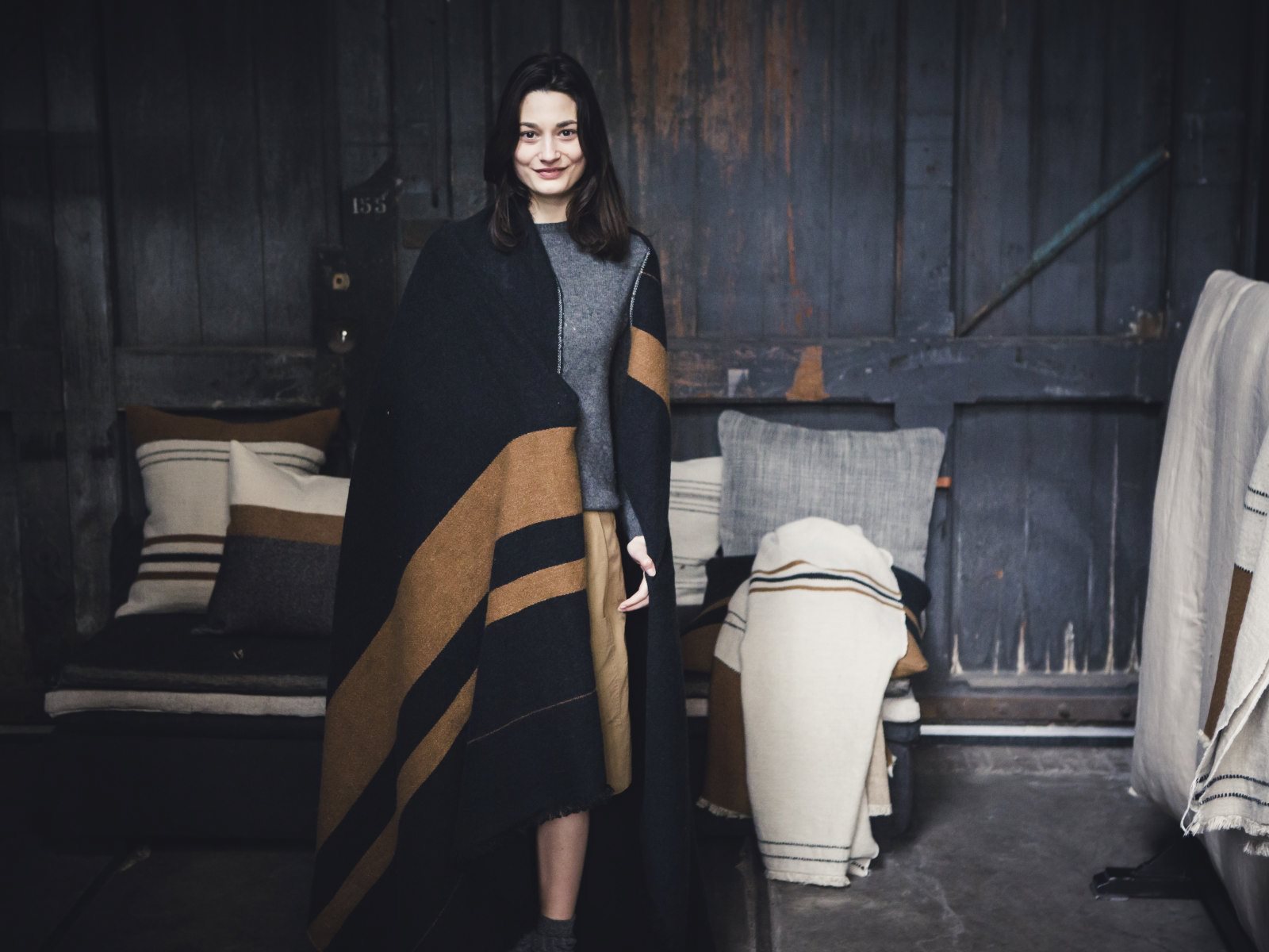 Eine lächelnde Frau steht drinnen, eingewickelt in die Oscar Leinen-Wolle-Mix von Libeco-Decke mit braunen Streifen. Hinter ihr rahmt eine rustikale Holzwand eine gemütliche Szene mit einem Sofa, Kissen und gefalteten Decken ein.