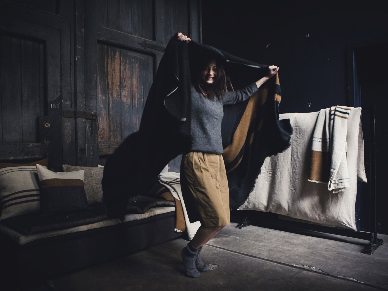 Eine Person in einem grauen Pullover und einem hellbraunen Rock steht in einem Innenraum auf Beton und hält spielerisch die Decke Oscar Leinen-Wolle-Mix von Libeco. Dahinter stehen ein Sofa und ein Regal mit gefalteten und aufgehängten gestreiften Decken.