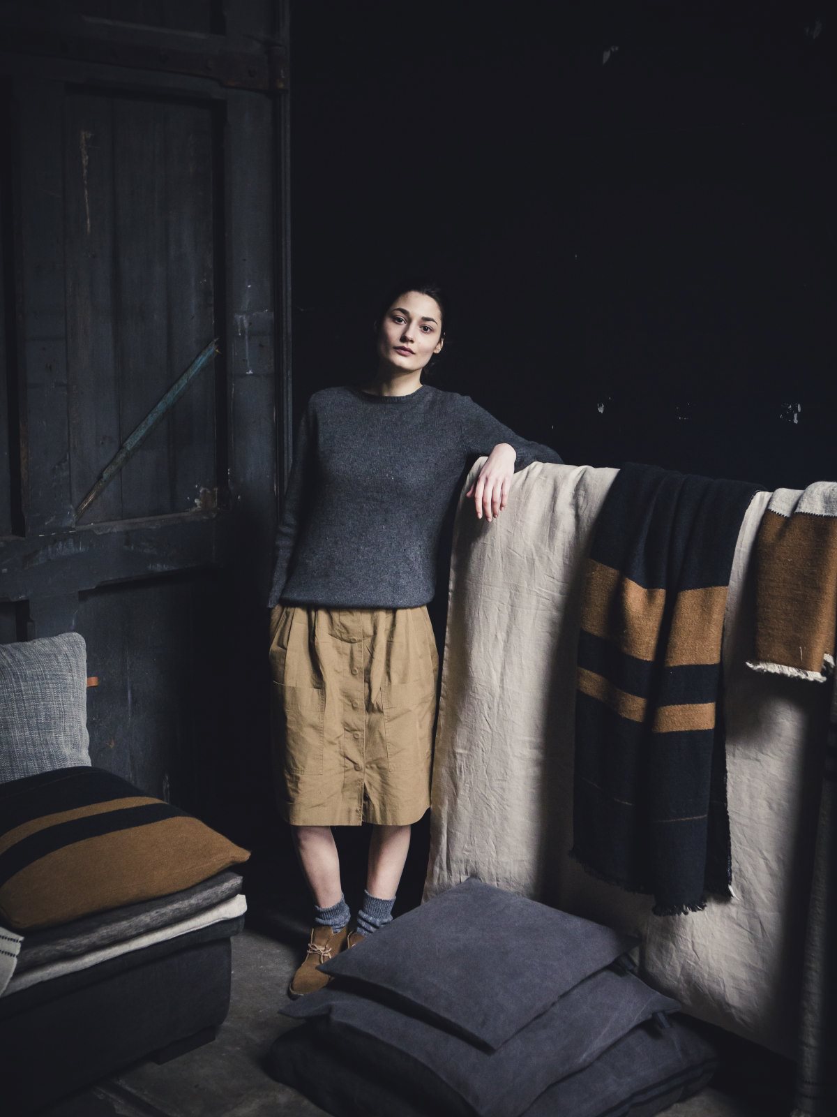 Eine Frau in grauem Pullover und hellbraunem Rock lehnt auf einer Stofffläche, umgeben von Oscar Leinen-Wolle-Mix von Libeco-Decken und -Kissen in einem schummrigen, rustikalen Raum mit dunklen Wänden und Holzpaneelen.