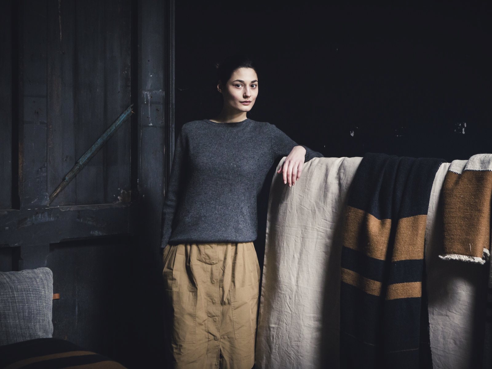 Eine Frau in einem grauen Pullover und einer hellbraunen Hose lehnt an einem Oscar Leinen-Wolle-Mix von Libeco, der über eine Schiene in einem schwach beleuchteten, rustikalen Raum mit dunklen Wänden drapiert ist.