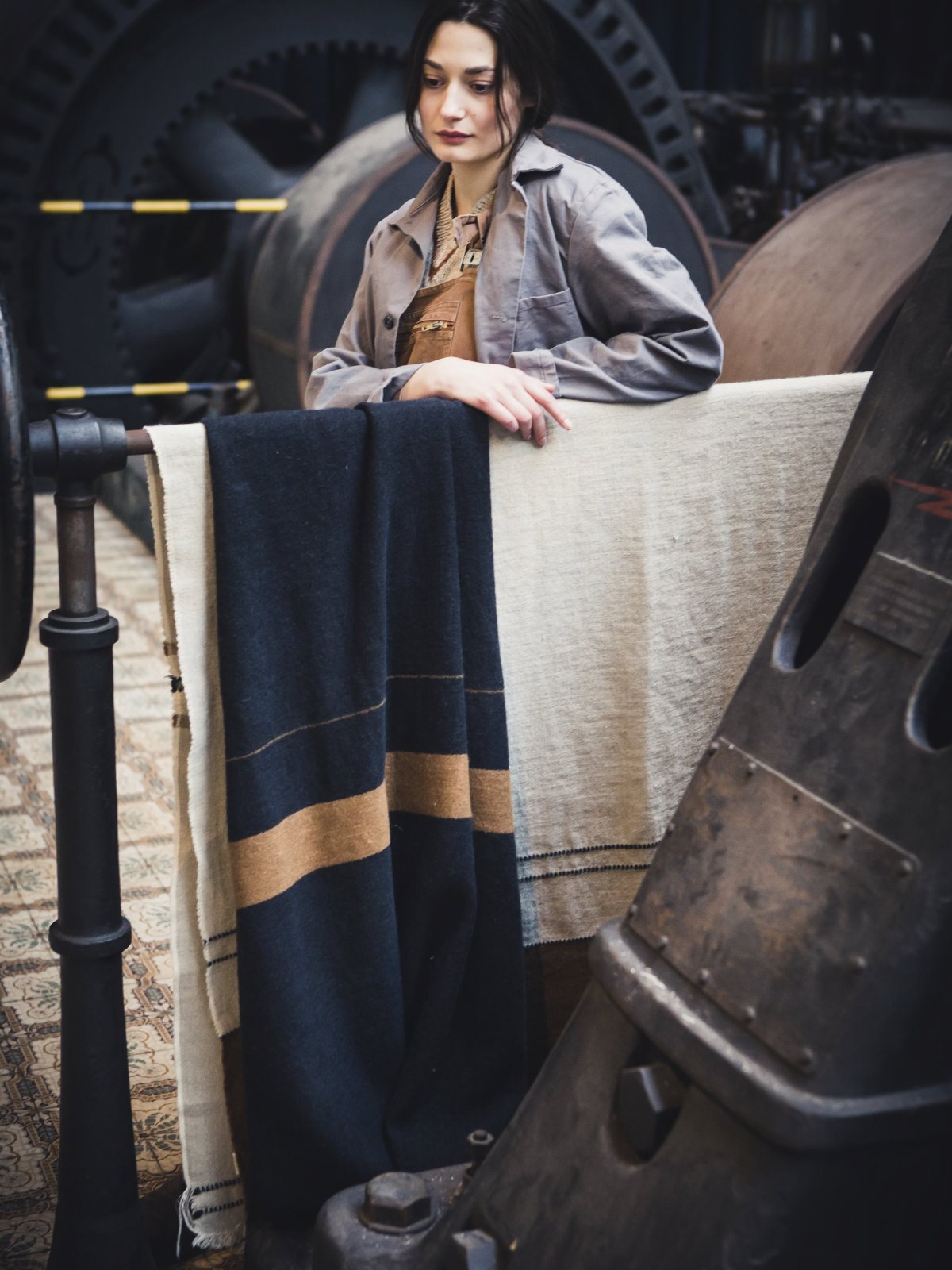 Eine Frau in grauer Arbeitsjacke und brauner Latzhose lehnt an einem Geländer, das mit Oscar Leinen-Wolle-Mix von Libeco-Decken bedeckt ist, in einer Industrieumgebung mit großen Maschinen und Metallrohren.