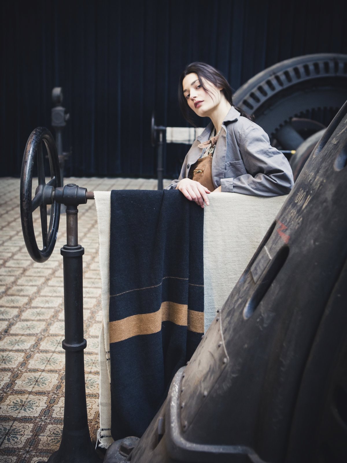 Eine Frau im grauen Hemd lehnt an einer großen Maschine, über ein Metallgeländer sind Oscar Leinen-Wolle-Mix von Libeco-Decken drapiert. Der Raum hat dunkle Wände und einen gemusterten Kachelboden.