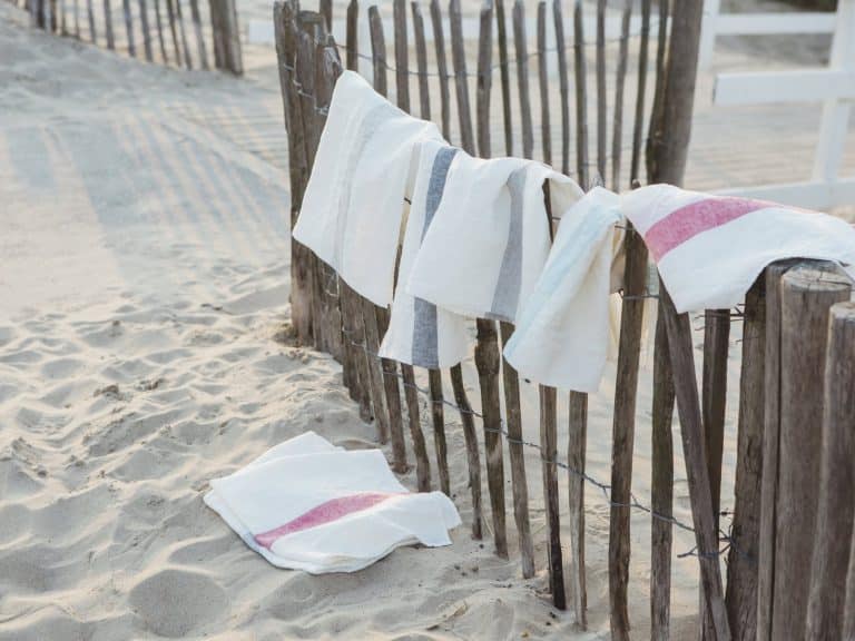 Weiße Handtücher mit farbigen Streifen, darunter die Outlast und Maize Kissen von HEFEL, hängen an einem rustikalen Holzzaun am Strand, wobei ein Handtuch im Sand liegt - so entsteht eine entspannte Küstenatmosphäre.
