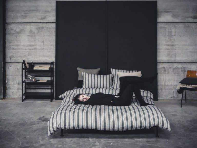 A person dressed in black lies on a striped bed made of The Tack Stripe linen bedding from Libeco in an industrial-style room with concrete walls, minimalist furniture and a bookshelf.