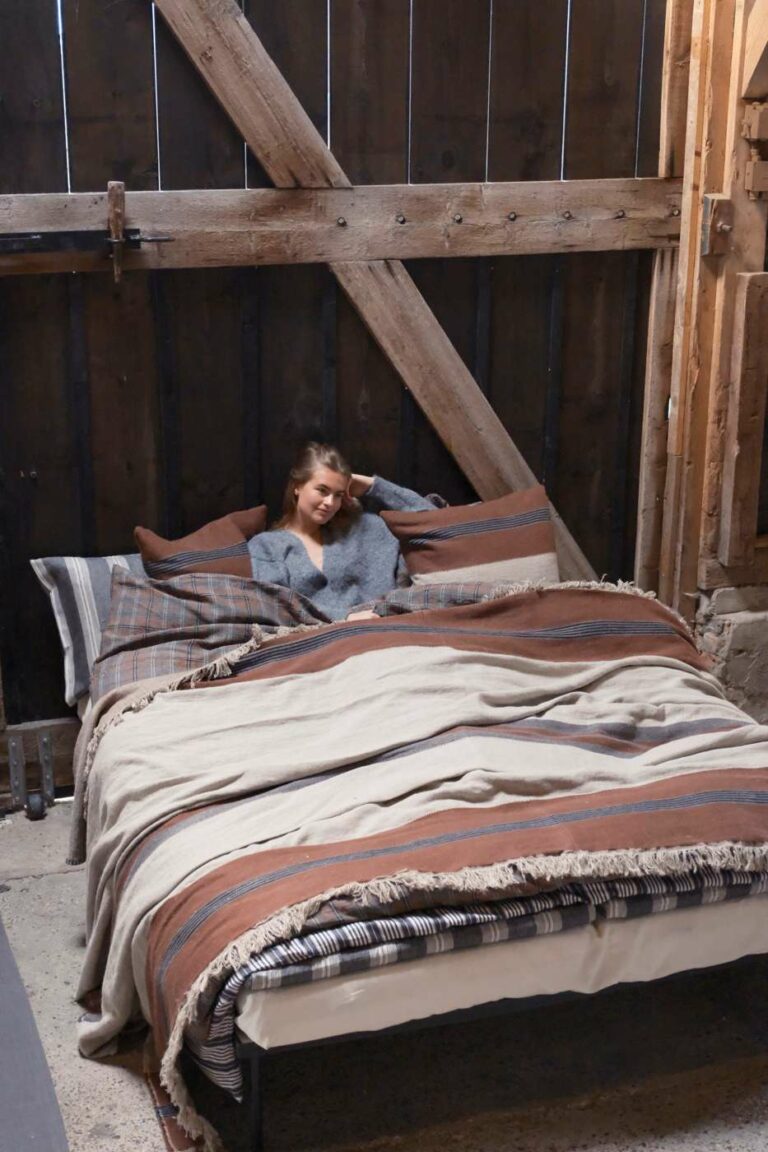 Eine Frau sitzt lächelnd im Bett und stützt den Kopf auf ihre Hand, eingewickelt in kuschelige Sienna Leinen und Wolle-Mix von Libeco Decken, in einem rustikalen Zimmer mit Holzwänden und Balken.