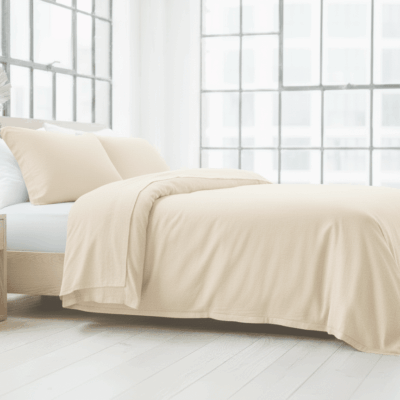 A modern bedroom with a large bed covered with hemp and linen bedding Oyster by Vieböck 226/02, two pillows, a wooden bedside table with a white vase and large windows that fill the bright, minimalist room with natural sunlight.