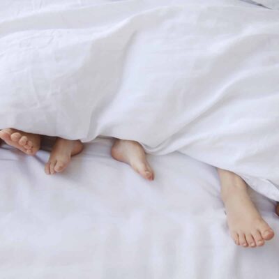 Four pairs of feet peek out from under a white blanket on a bed, suggesting that several people are lying together and relaxing. The white bedding creates a soft and cozy atmosphere.