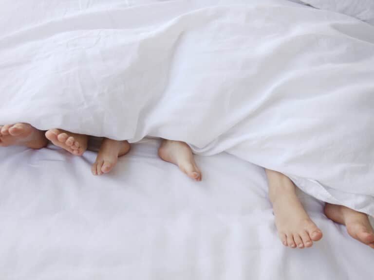 Four pairs of feet peek out from under a white blanket on a bed, suggesting that several people are lying together and relaxing. The white bedding creates a soft and cozy atmosphere.