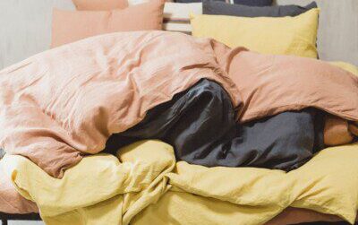 Ein unordentliches, ungemachtes Bett mit orangefarbener, gelber und grauer Bettwäsche - darunter Madison Navy Farbige Leinen Bettwäsche von Libeco - und mehreren Kissen vor einer hellen Wand.