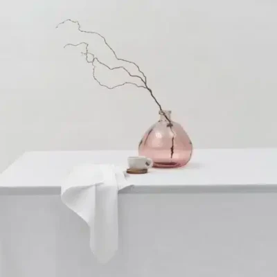 Eine rosafarbene Glasvase mit dünnen Zweigen, eine weiße Tasse mit Untertasse und eine gefaltete weiße Serviette stehen auf einem weißen Tisch mit einer weißen Tischdecke vor einem schlichten weißen Hintergrund.