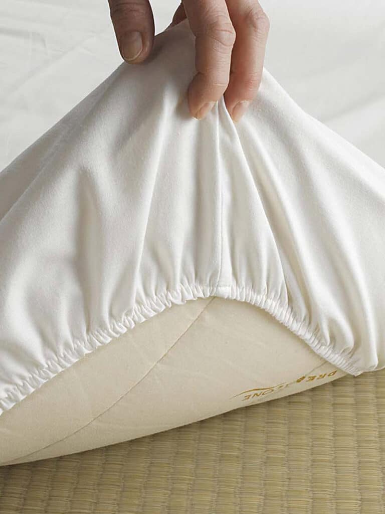 Eine Hand spannt den elastischen Rand eines weißen Spannbetttuchs über die Ecke einer Matratze. Die Oberfläche darunter ist eine gewebte Matte.